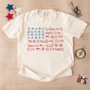 May include: Cream-colored t-shirt featuring a unique American flag design. The flag's stars are blue starfish, and the stripes are composed of red sea creatures. The shirt is a comfortable, casual wear item.