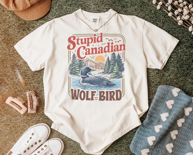 May include: Cream-colored t-shirt with a graphic that reads "Stupid Canadian Wolf Bird." The graphic features a loon on a lake with trees and a sunset. The shirt is styled with a necklace, shoes, and a hat.