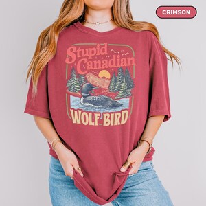 May include: Crimson-colored t-shirt with a graphic design featuring a loon on a lake, trees, and the text "Stupid Canadian Wolf Bird". The design has a vintage, outdoorsy aesthetic.