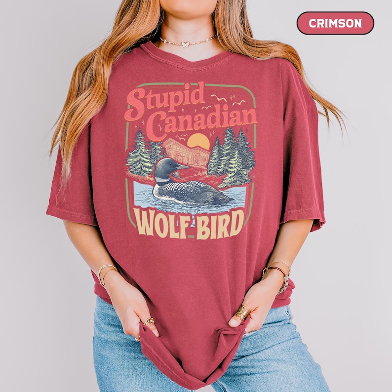 May include: Crimson-colored t-shirt with a graphic design featuring a loon on a lake, trees, and the text "Stupid Canadian Wolf Bird". The design has a vintage, outdoorsy aesthetic.