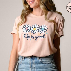 May include: A peach-colored t-shirt featuring a daisy graphic with pink, yellow, and blue accents. The phrase "life is good" is written in a cursive font below the flowers. The shirt is made of soft material.