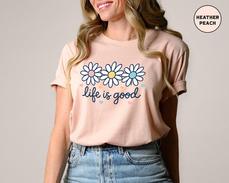 May include: A peach-colored t-shirt featuring a daisy graphic with pink, yellow, and blue accents. The phrase "life is good" is written in a cursive font below the flowers. The shirt is made of soft material.
