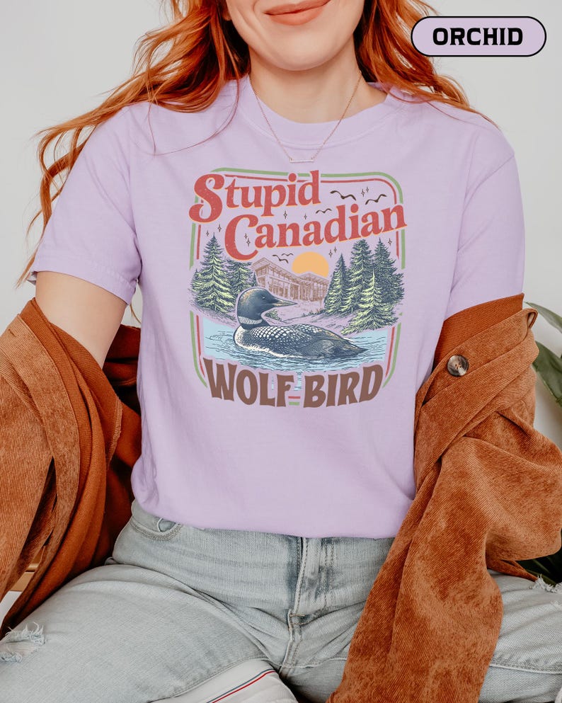 May include: Orchid-colored t-shirt with a graphic that reads "Stupid Canadian Wolf Bird." The design features a loon in a lake, trees, and a sunset. The shirt is short-sleeved and has a crew neck.