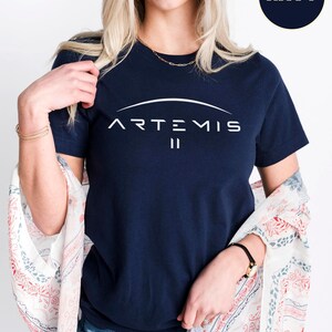 May include: Navy blue t-shirt with the word "ARTEMIS" in white, arched above the Roman numeral "II". The shirt is a crew neck style. A small circle in the upper right corner reads "NAVY".