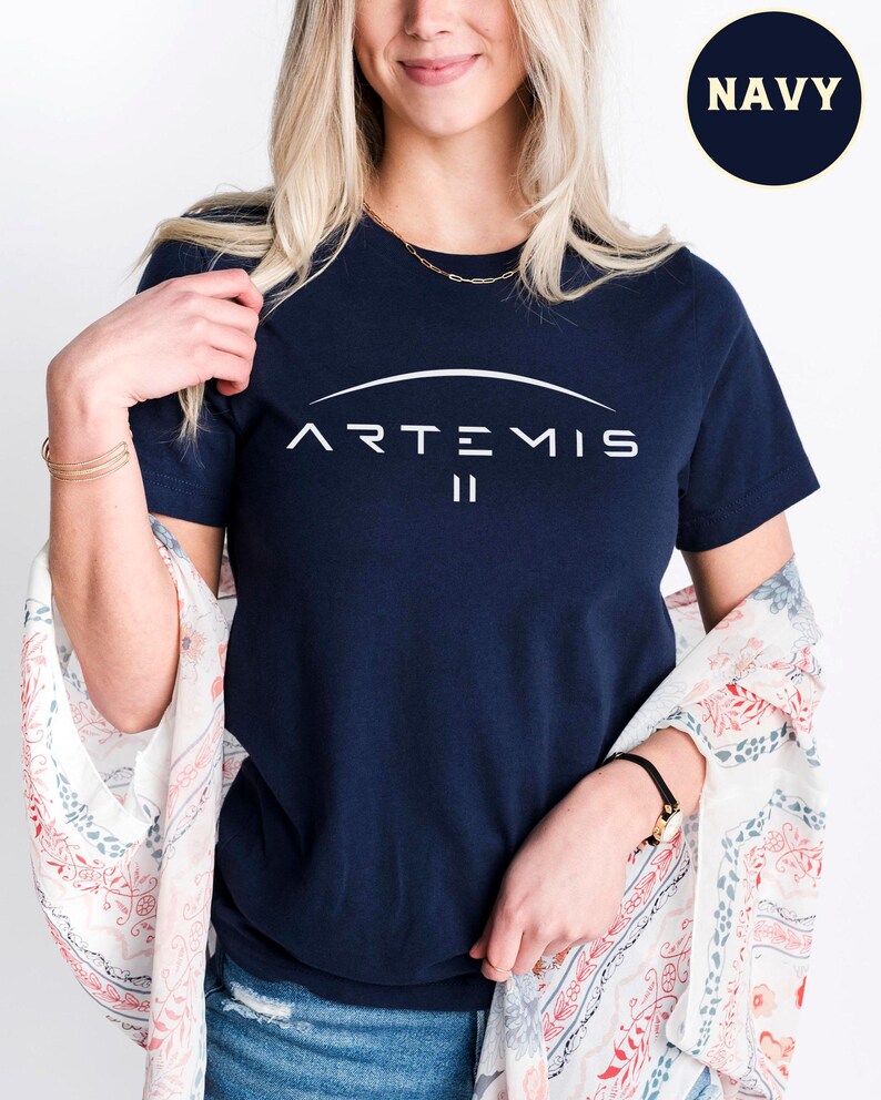 May include: Navy blue t-shirt with the word "ARTEMIS" in white, arched above the Roman numeral "II". The shirt is a crew neck style. A small circle in the upper right corner reads "NAVY".