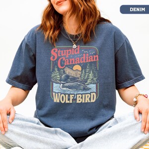 May include: Denim blue t-shirt with a graphic featuring a loon on a lake, trees, and the text "Stupid Canadian WOLF BIRD". The design has a vintage, outdoorsy feel. The shirt is worn by a person.