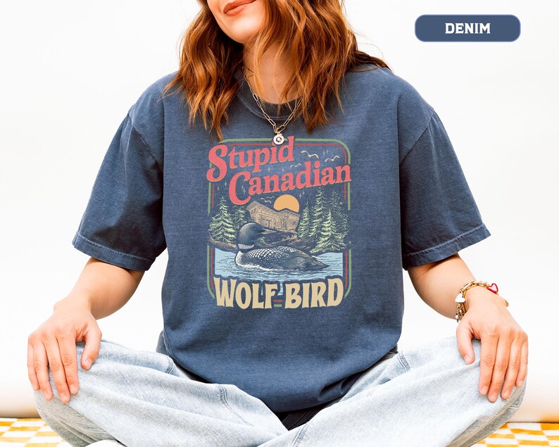 May include: Denim blue t-shirt with a graphic featuring a loon on a lake, trees, and the text "Stupid Canadian WOLF BIRD". The design has a vintage, outdoorsy feel. The shirt is worn by a person.