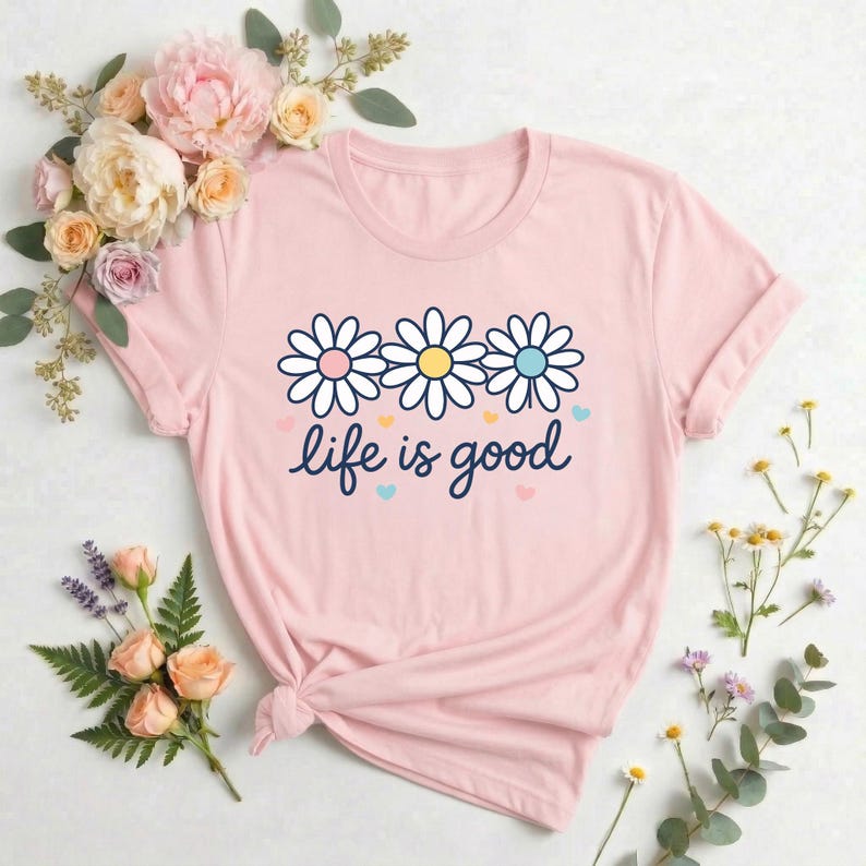 May include: A light pink t-shirt featuring a daisy graphic with pink, yellow, and blue centers, and the phrase "life is good" in cursive. The shirt is surrounded by flowers.