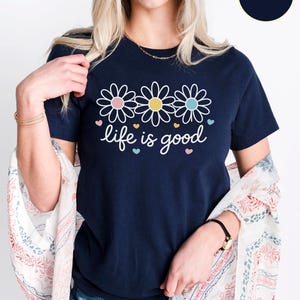 May include: Navy blue t-shirt featuring three daisy flowers with pink, yellow, and blue centers, and the phrase "life is good" in white script. The shirt is a casual, everyday wear item.