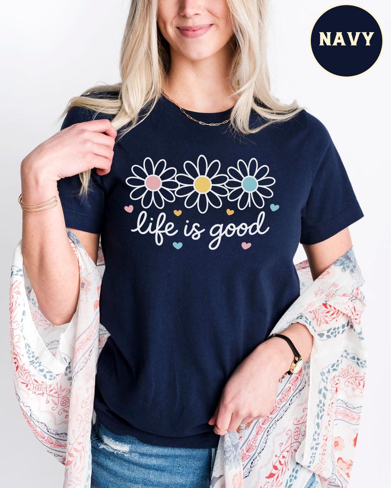 May include: Navy blue t-shirt featuring three daisy flowers with pink, yellow, and blue centers, and the phrase "life is good" in white script. The shirt is a casual, everyday wear item.