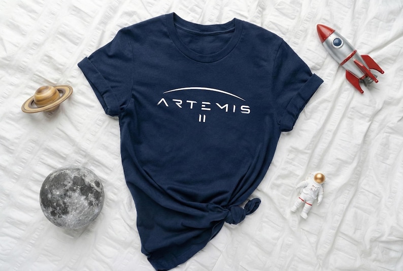 May include: Navy blue t-shirt with the white text "ARTEMIS II" in a curved design. The shirt is surrounded by space-themed toys, including a rocket, astronaut, Saturn, and a moon.