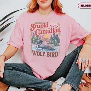 May include: A light pink t-shirt with the text "Stupid Canadian Wolf Bird" in a retro design. The graphic features a loon on a lake with trees and a sunset. The shirt is a relaxed fit.