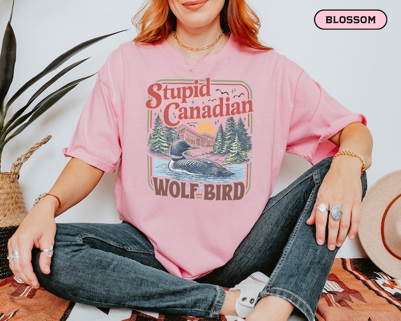 May include: A light pink t-shirt with the text "Stupid Canadian Wolf Bird" in a retro design. The graphic features a loon on a lake with trees and a sunset. The shirt is a relaxed fit.
