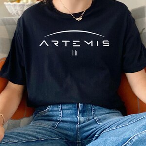 May include: Black t-shirt with the word "ARTEMIS" in white, arched above the Roman numeral "II". The shirt is short-sleeved and made of a soft material. The person is wearing blue jeans.