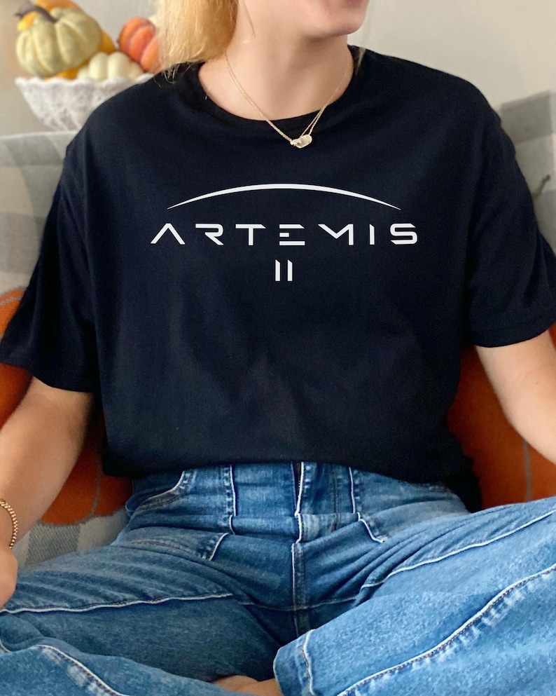 May include: Black t-shirt with the word "ARTEMIS" in white, arched above the Roman numeral "II". The shirt is short-sleeved and made of a soft material. The person is wearing blue jeans.