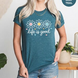 May include: Teal t-shirt with the phrase "life is good" in white script, decorated with three daisy flowers and small hearts. The shirt has short sleeves and a round neckline. The text "Heather Deep Teal" is in a circle in the upper right corner.