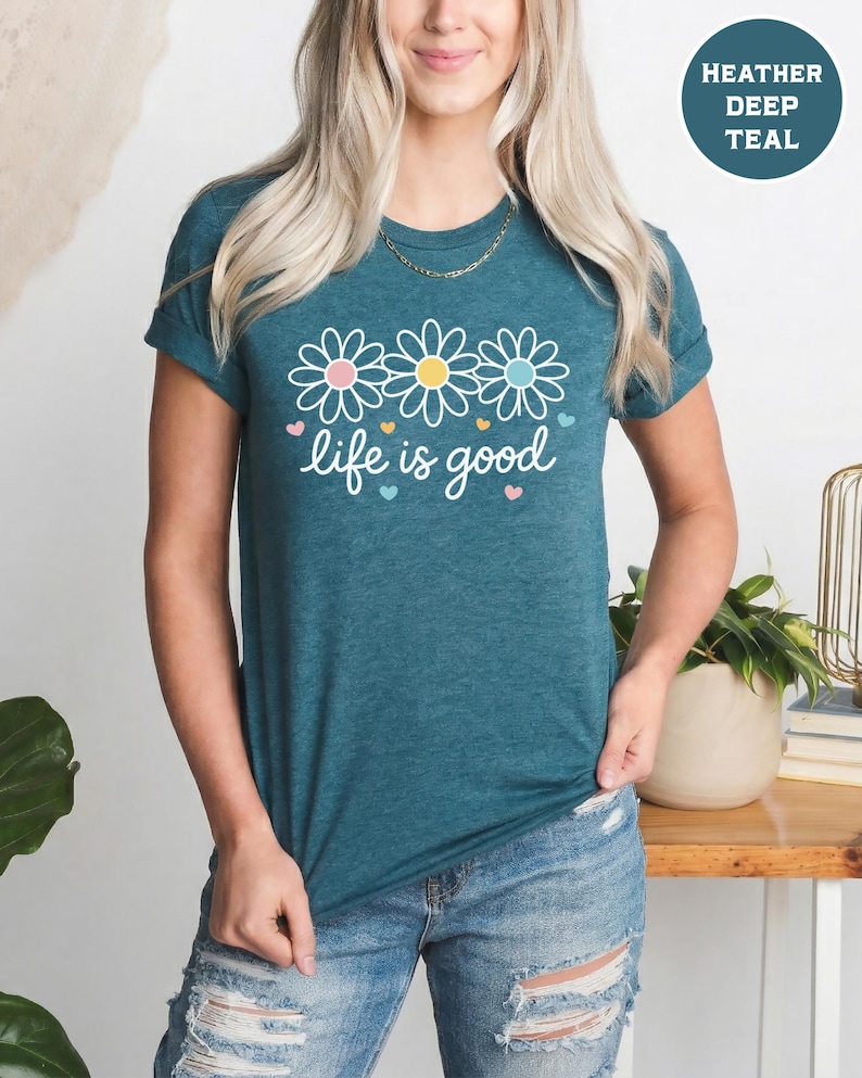 May include: Teal t-shirt with the phrase "life is good" in white script, decorated with three daisy flowers and small hearts. The shirt has short sleeves and a round neckline. The text "Heather Deep Teal" is in a circle in the upper right corner.