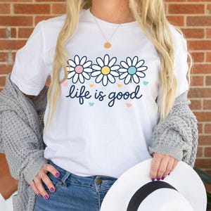 May include: White t-shirt with a floral design and the phrase "life is good" in a script font. The shirt features three daisy-like flowers with pink, yellow, and blue centers. Small heart shapes in pastel colors are scattered around the text.