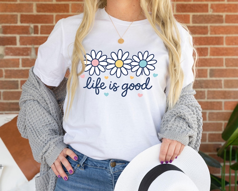 May include: White t-shirt with a floral design and the phrase "life is good" in a script font. The shirt features three daisy-like flowers with pink, yellow, and blue centers. Small heart shapes in pastel colors are scattered around the text.