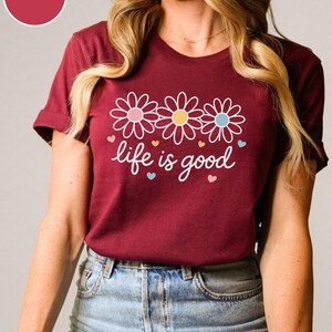 May include: Maroon t-shirt with a floral design and the phrase "life is good" in white script. The design features three daisies with pink, yellow, and blue centers, and small heart accents. The shirt is made of a soft material.