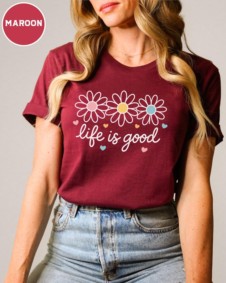 May include: Maroon t-shirt with a floral design and the phrase "life is good" in white script. The design features three daisies with pink, yellow, and blue centers, and small heart accents. The shirt is made of a soft material.