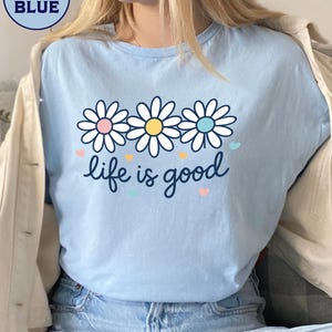 May include: Light blue t-shirt with a daisy graphic and the phrase "life is good." The daisy design features white petals with pink, yellow, and blue centers. Small hearts in various colors are scattered around the text.