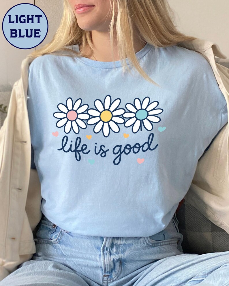 May include: Light blue t-shirt with a daisy graphic and the phrase "life is good." The daisy design features white petals with pink, yellow, and blue centers. Small hearts in various colors are scattered around the text.