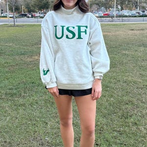 Könnte beinhalten: Ein hellgraues Sweatshirt mit den Buchstaben "USF" in Grün. Das Sweatshirt hat einen hohen Kragen und lange Ärmel. Die Person trägt schwarze Shorts und weiße Turnschuhe. Der Hintergrund ist eine grasbewachsene Fläche mit Bäumen und einem bewölkten Himmel.