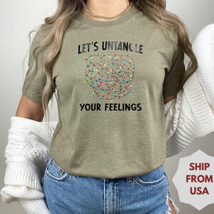 May include: Olive green t-shirt with the text "LET'S UNTANGLE YOUR FEELINGS" above a graphic of tangled Christmas lights. The shirt is made of a soft material and is suitable for casual wear.