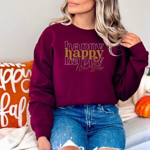 May include: Burgundy sweatshirt with the words "happy happy happy New Year" in a layered design. The word "happy" is in gold, and the other words are in white. The model is wearing blue denim shorts.