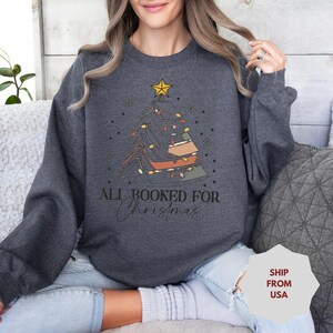 May include: Dark gray sweatshirt with a Christmas tree design. The tree is decorated with lights and a star on top. The text "ALL BOOKED FOR Christmas" is printed below the tree.
