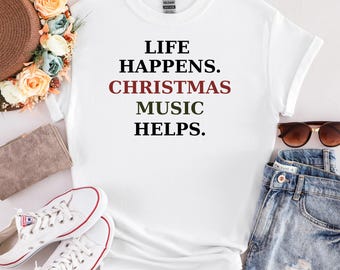 May include: White t-shirt with the text "LIFE HAPPENS. CHRISTMAS MUSIC HELPS." in black, red, and green. The shirt is displayed with sunglasses, denim shorts, and white sneakers.