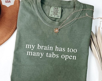 May include: Olive green t-shirt with the phrase "my brain has too many tabs open" in white. A gold necklace with a pendant rests on the shirt. The shirt is folded and has a Comfort Colors tag.