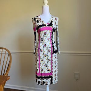 May include: A long-sleeve knee-length dress featuring a cream base with a vibrant pattern of pink panels, black borders, and small floral motifs. The garment has a V-neckline and a fitted silhouette, displayed on a white mannequin torso.