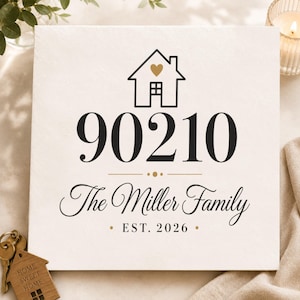 May include: A square white sign featuring a black house icon with a gold heart, the number 90210 in black, and the text The Miller Family in black script. Below is the text EST. 2026 with gold dots. A wooden house-shaped keychain is visible nearby.