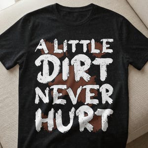 Könnte beinhalten: Dunkelgraues T-Shirt mit dem weißen Text "A Little Dirt Never Hurt" und einem braunen, schmutzähnlichen Design. Das T-Shirt hat kurze Ärmel und einen Rundhalsausschnitt.