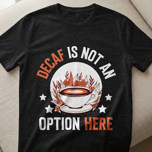 Könnte beinhalten: Schwarzes T-Shirt mit einem Kaffee-Motiv. Das Design zeigt eine weiße Kaffeetasse mit brauner Flüssigkeit, umgeben von Blumen und dem Text "DECAF IS NOT AN OPTION HERE" in Orange und Weiß.