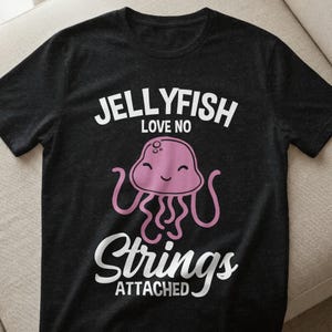 Könnte beinhalten: Schwarzes T-Shirt mit einem rosa Quallen-Motiv und dem weißen Text "JELLYFISH LOVE NO STRINGS ATTACHED". Die Qualle hat ein lächelndes Gesicht und wellenförmige Tentakel. Ein lässiges, lustiges Design.