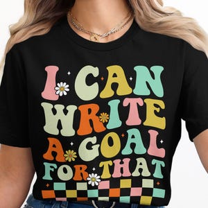 Könnte beinhalten: Schwarzes T-Shirt mit dem Text "I CAN WRITE A GOAL FOR THAT" in farbenfrohen Buchstaben im Retro-Stil. Das Design beinhaltet Gänseblümchen und ein Schachbrettmuster. Ein lässiges, motivierendes T-Shirt.
