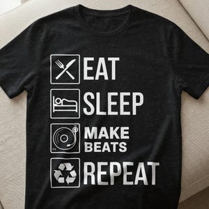 May include: Black t-shirt with white text and graphics. The text reads "EAT SLEEP MAKE BEATS REPEAT" with corresponding icons: a fork and knife, a bed, a record player, and a recycling symbol. The shirt is made of a soft material.