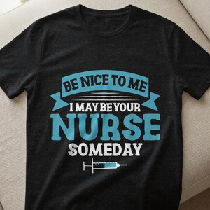 Könnte beinhalten: Dunkelgraues T-Shirt mit dem Text "BE NICE TO ME I MAY BE YOUR NURSE SOMEDAY" in Blau und Weiß. Eine Spritzengrafik befindet sich darunter. Das T-Shirt hat einen Rundhalsausschnitt.