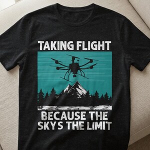 Könnte beinhalten: Schwarzes T-Shirt mit einem Grafikdesign, das eine Drohne, Berge und Bäume zeigt. Der Text "TAKING FLIGHT" steht über der Drohne und "BECAUSE THE SKY'S THE LIMIT" darunter. Das Design ist in Weiß und Türkis gehalten.