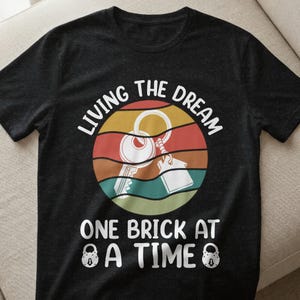 Könnte beinhalten: Schwarzes T-Shirt mit einem grafischen Design, das einen Schlüssel und eine Haussilhouette in einem kreisförmigen Sonnenuntergangsdesign zeigt. Der Text lautet "LIVING THE DREAM" und "ONE BRICK AT A TIME", mit Vorhängeschloss-Symbolen.