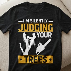 Op de afbeelding: Zwart t-shirt met de tekst "I'M SILENTLY JUDGING YOUR TREES" in wit en geel. De afbeelding bevat een boom, een persoon en een kettingzaag, allemaal in wit.