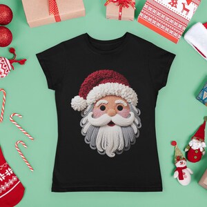 Könnte beinhalten: Schwarzes T-Shirt mit einem Weihnachtsmann-Gesichtsmotiv. Das Gesicht des Weihnachtsmanns hat einen roten Hut, einen weißen Bart und rosige Wangen. Weihnachtsdekorationen, darunter Kugeln, Geschenke und Zuckerstangen, umgeben das T-Shirt.