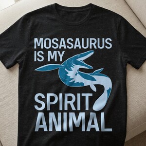 Op de afbeelding: Donkergrijs t-shirt met een blauwe mosasaurus illustratie en de tekst "MOSASAURUS IS MY SPIRIT ANIMAL" in lichtblauwe letters. Het ontwerp staat in het midden van de voorkant.