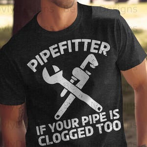 Può includere: T-shirt grigio scuro con la scritta bianca "PIPEFITTER" e chiavi incrociate. Sotto, la frase bianca "IF YOUR PIPE IS CLOGGED TOO". La maglietta è a girocollo.