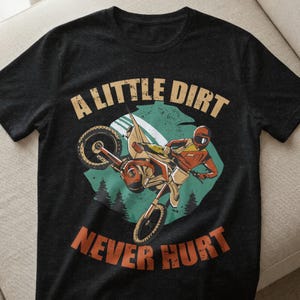Può includere: T-shirt grigio scuro con grafica motocross. Il design presenta un pilota su una moto da cross a mezz'aria, con la scritta "A LITTLE DIRT NEVER HURT" in un font invecchiato. La grafica include sfumature di arancione, beige e verde acqua.