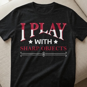 Könnte beinhalten: Dunkelgraues T-Shirt mit dem Text "I PLAY WITH SHARP OBJECTS" in Rot und Weiß. Der Text ist mittig auf dem Shirt platziert, mit dekorativen Elementen über und unter den Worten.