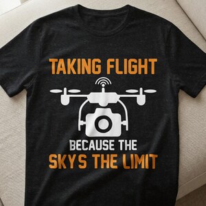 Puede incluir: Camiseta negra con un gráfico de dron blanco y el texto naranja "TAKING FLIGHT BECAUSE THE SKYS THE LIMIT". El gráfico del dron incluye una lente de cámara y una antena.